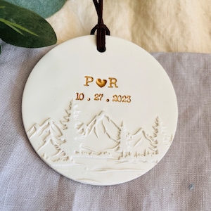 Może przedstawiać: Biała ceramiczna ozdoba z motywem g&oacute;rskim i napisem "P &hearts; R 10.27.2023" w kolorze złotym.