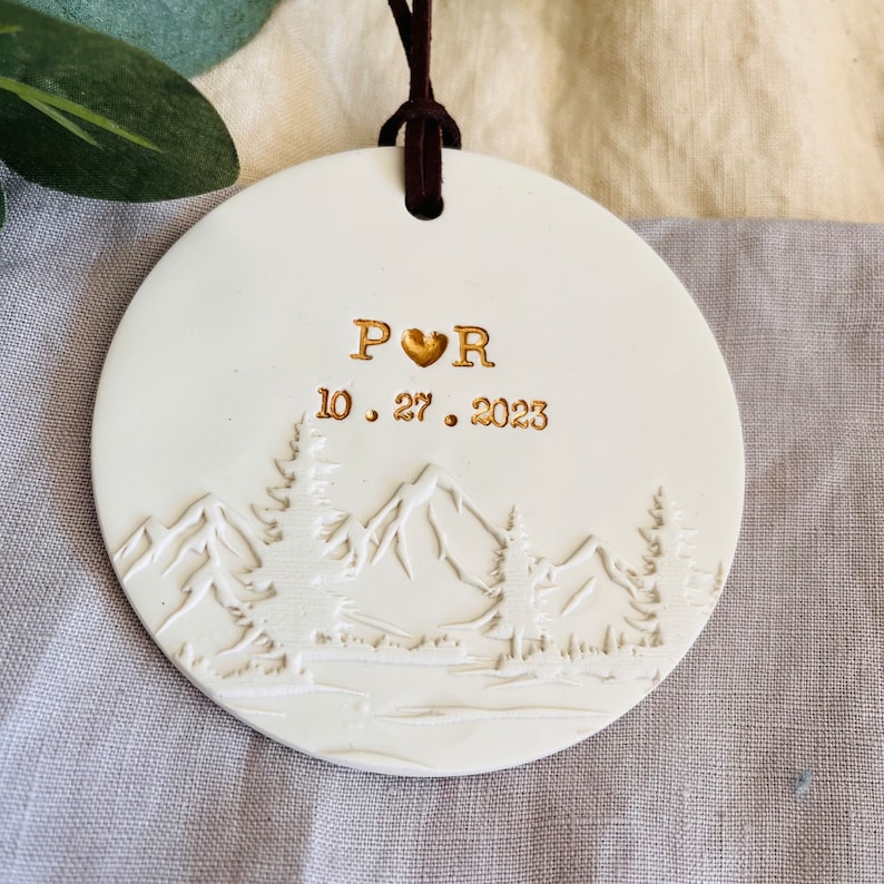 Może przedstawiać: Biała ceramiczna ozdoba z motywem g&oacute;rskim i napisem "P &hearts; R 10.27.2023" w kolorze złotym.