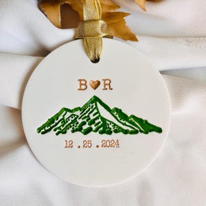 May include: White ceramic bauble featuring a green mountain range design, the letters "B & R" with a heart in between, and the date "12.25.2024" in copper-coloured lettering.