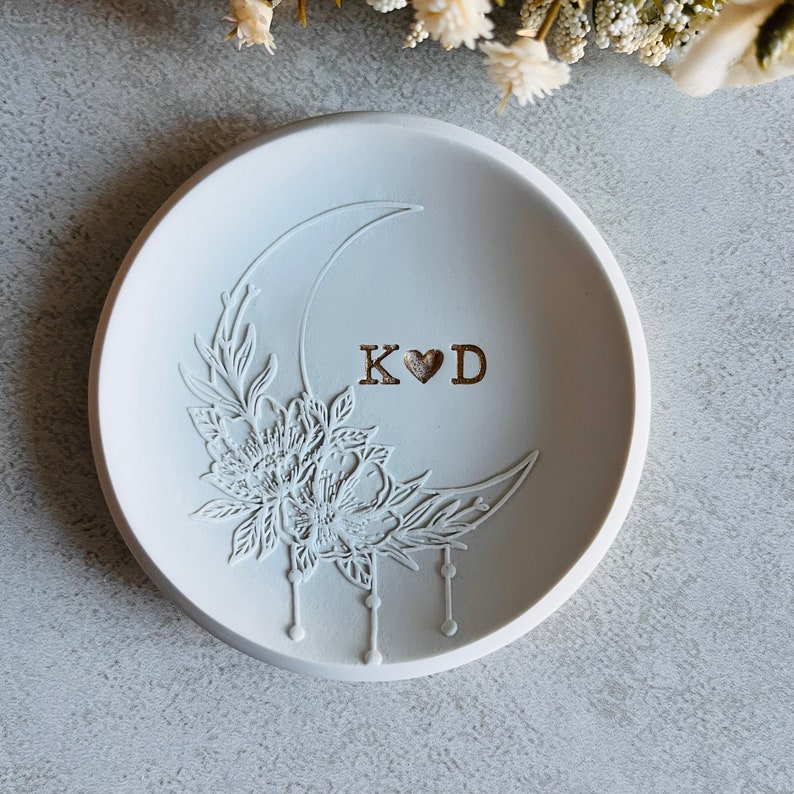 Puede incluir: Plato para anillos de cer&aacute;mica blanca con un dise&ntilde;o de media luna y detalles florales. El plato est&aacute; grabado con las iniciales "K&hearts;D".