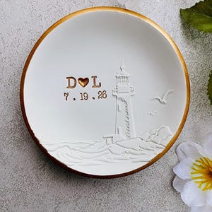 May include: A round white ceramic trinket dish with a gold-painted rim. The center features a raised white lighthouse relief, two flying birds, and gold-colored text reading D heart L and the date 7.19.26 embossed on the smooth surface.