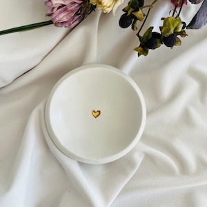 Könnte beinhalten: Weiße Keramik-Schmuckschale mit einem kleinen goldenen Herz in der Mitte.