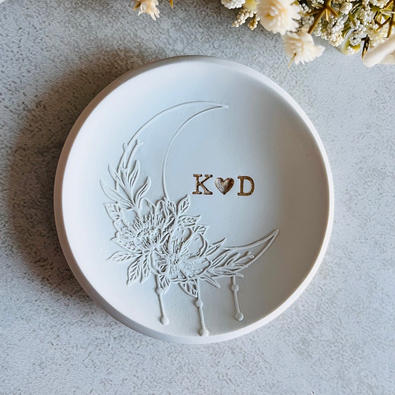 Puede incluir: Plato para anillos de cer&aacute;mica blanca con un dise&ntilde;o de luna creciente y detalles florales. El plato est&aacute; grabado con las iniciales "K&hearts;D".
