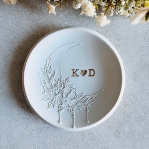 Puede incluir: Plato de cer&aacute;mica blanco para anillos con un dise&ntilde;o de media luna y detalles florales. El plato est&aacute; grabado con las iniciales "K&hearts;D".