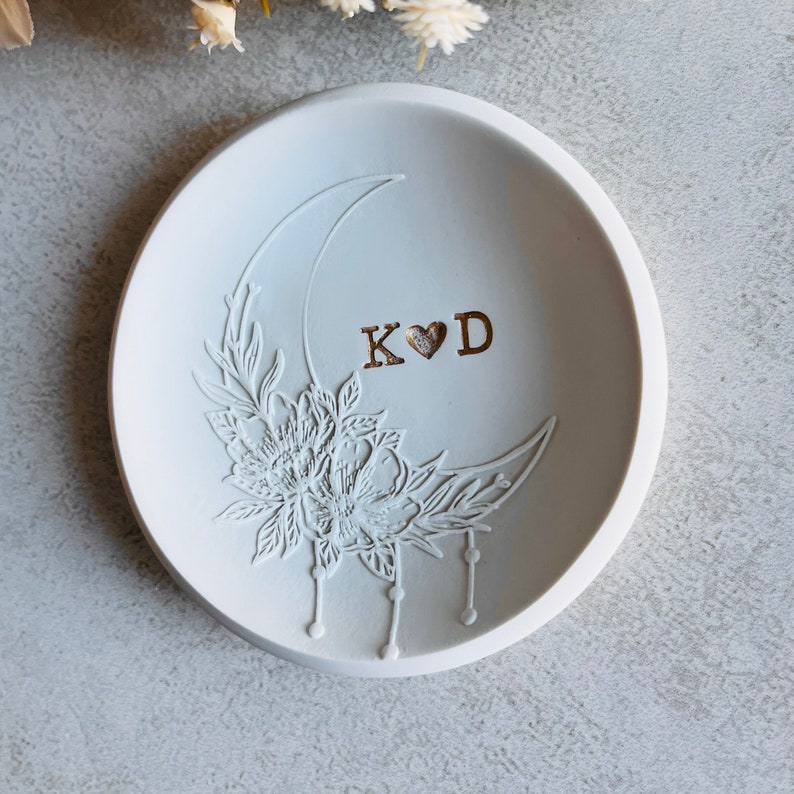 Puede incluir: Un porta anillos de cer&aacute;mica blanca con un dise&ntilde;o de luna creciente y detalles florales. El plato est&aacute; grabado con las iniciales "K&hearts;D" en oro.