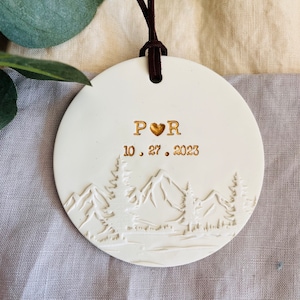 Może przedstawiać: Biała ozdoba ceramiczna z motywem g&oacute;rskim, z inicjałami "P♡R" i datą "10.27.2023" w złotych literach.