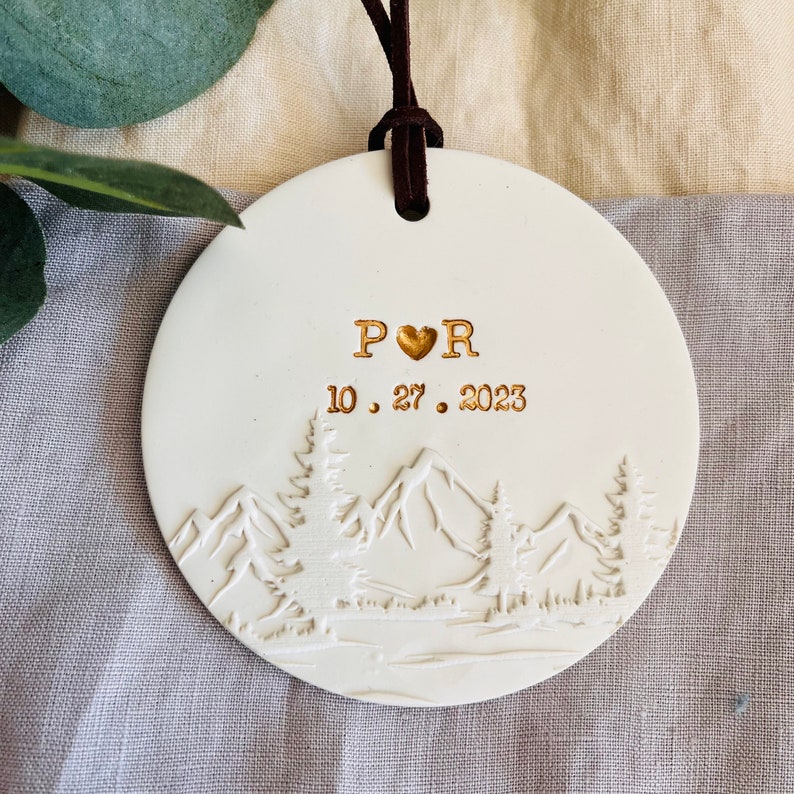 Może przedstawiać: Biała ozdoba ceramiczna z motywem g&oacute;rskim, z inicjałami "P♡R" i datą "10.27.2023" w złotych literach.