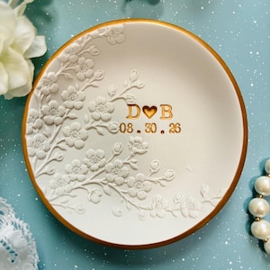 May include: A round white ceramic trinket dish with a gold-painted rim. The surface features a raised, embossed floral branch design on the left side. The center displays the gold-colored text D heart B and the date 08.30.26 in a serif font.