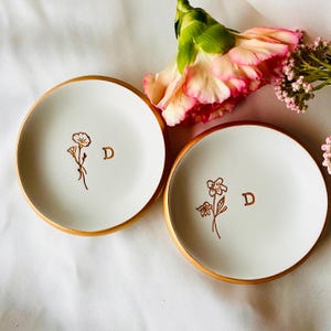 Puede incluir: Dos platos de anillo de cerámica blanca con bordes dorados. Cada plato presenta un dibujo simple de una flor y la letra "D" en oro.