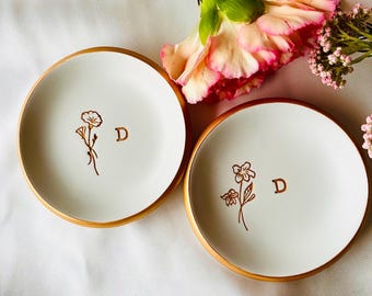 Può includere: Due porta anelli in ceramica bianca con bordi dorati. Ogni piatto presenta un semplice disegno di un fiore e la lettera "D" in oro.