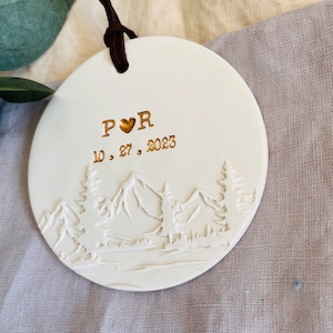 Może przedstawiać: Biała ozdoba ceramiczna z motywem g&oacute;r w kolorze złotym i tekstem "P &hearts; R 10. 27. 2023"