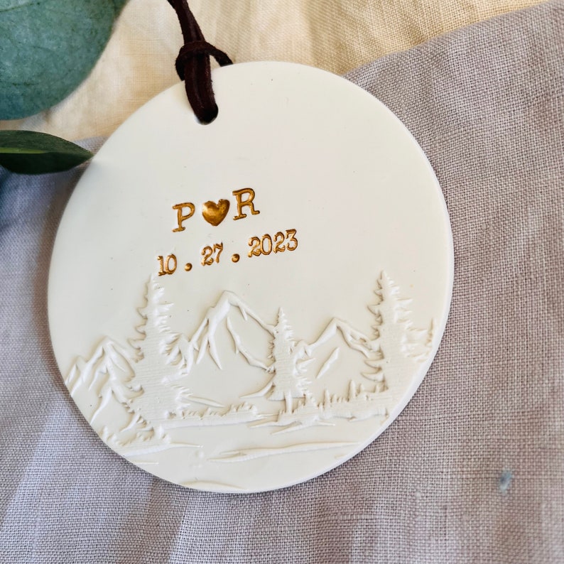 Może przedstawiać: Biała ozdoba ceramiczna z motywem g&oacute;r w kolorze złotym i tekstem "P &hearts; R 10. 27. 2023"