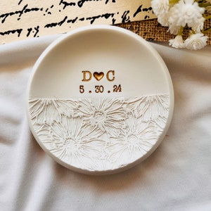 May include: White ceramic ring dish with a floral design and gold lettering. The lettering reads "D ♥ C 5.30.24".