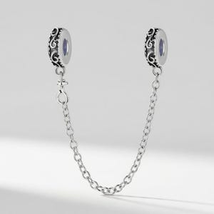 May include: A silver-colored charm bracelet safety chain with two decorative beads. Each bead has an ornate, swirling design and a small, oval-shaped opening. A delicate chain connects the beads.