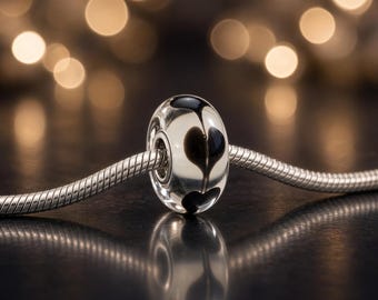 May include: A clear glass bead with a black heart pattern threaded onto a silver snake chain. The bead features a smooth, rounded shape with a central hole for the chain, showcasing a reflective surface and a distinct dark heart design.