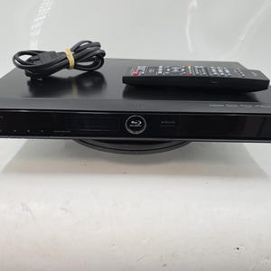 May include: A black Sharp Blu-ray player with a remote control and power cord. The player has a sleek design with a display screen and the Blu-ray logo. The remote control is black with multiple buttons.
