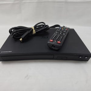 May include: A black Samsung Blu-ray disc player with a textured, ribbed top surface. The device includes a black remote control with numerous buttons and a coiled black power cord secured with a tan rubber band, all resting on a white surface.