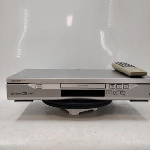 May include: A silver Sharp DVD player with a rectangular chassis, a central disc tray, and a front control panel featuring power, play, and stop buttons. A matching silver remote control rests on the top surface of the electronic device.