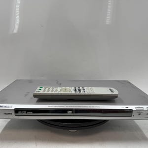 May include: Silver Sony DVD player with a remote control. The DVD player has a sleek design with a front panel featuring the Sony logo, HDMI, and DVD symbols. The remote is light gray with multiple buttons.
