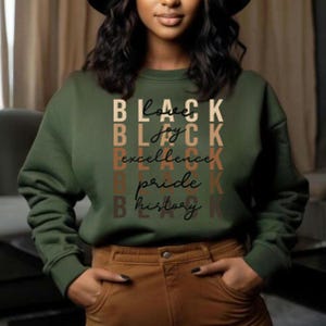 Może przedstawiać: Zielona bluza w kolorze oliwkowym z napisami "BLACK Love, joy, excellence, pride, history" w różnych czcionkach i kolorach. Bluza jest połączona z brązowymi spodniami. Osoba ma na sobie czarny kapelusz.