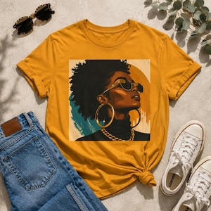 Op de afbeelding: Een mosterdgeel t-shirt met een grafische print van een persoon met een donkere huid en een groot afro-kapsel. De persoon draagt een donkere zonnebril, grote gouden oorringen en een gouden ketting tegen een cirkelvormige achtergrond.