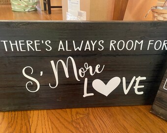 May include: A rustic, rectangular wooden sign with the phrase "THERE'S ALWAYS ROOM FOR S'MORE LOVE" in white lettering. The sign has a dark, stained finish and a white heart graphic.