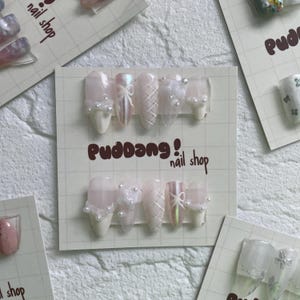May include: A set of six white and pink artificial nails with pearl and lace embellishments. The nails are displayed on a white card with the text "Puddoang! nail shop".