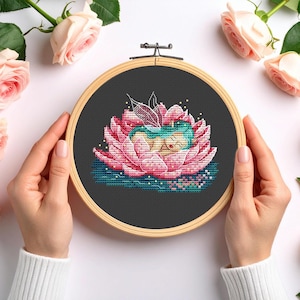 May include: A circular wooden embroidery hoop containing a cross-stitch design on black fabric. The design features a figure with teal hair and wings resting inside a large, multi-layered pink lotus flower, accented with small gold dots.