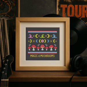 May include: A framed cross-stitch design featuring a dark gray background with colorful witch hats, moons, and mushrooms. The text "MAGIC & MUSHROOMS" is stitched in yellow. The frame is made of light brown wood.