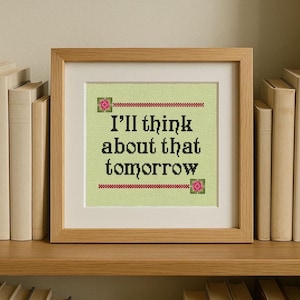 以下が含まれることがあります： 「I'll think about that tomorrow」のテキストが黒で、薄緑色の背景にクロスステッチされた額入りのアートワーク。明るい木製のフレームに入っており、本棚に飾られています。