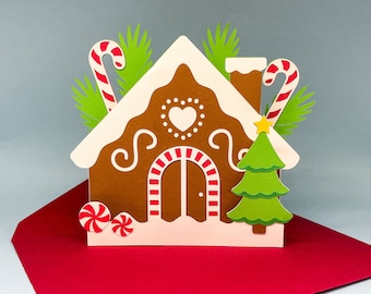 Könnte beinhalten: Eine dreidimensionale Grußkarte aus Papier in Form eines Lebkuchenhauses mit brauner Struktur, weißen Zuckergussdetails, einem Herzausschnitt, rot-weißen Zuckerstangen, grünen Tannenzweigen, einem kleinen grünen Weihnachtsbaum und zwei Pfefferminzbonbons.