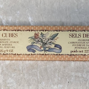 May include: A rectangular box with the words "BATH CUBES" and "SELS DE BAIN" in a decorative frame. The box is beige with a repeating pattern of small dots. The text includes ingredients and net weight of 225g.