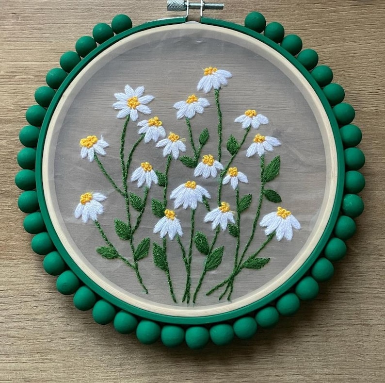 Puede incluir: Pieza de arte de aro floral bordado con margaritas blancas con centros amarillos y tallos y hojas verdes. El bordado est&aacute; sobre una tela transparente dentro de un aro de madera, rodeado por un borde de cuentas verdes. El aro mide unos 20 cm de di&aacute;metro.