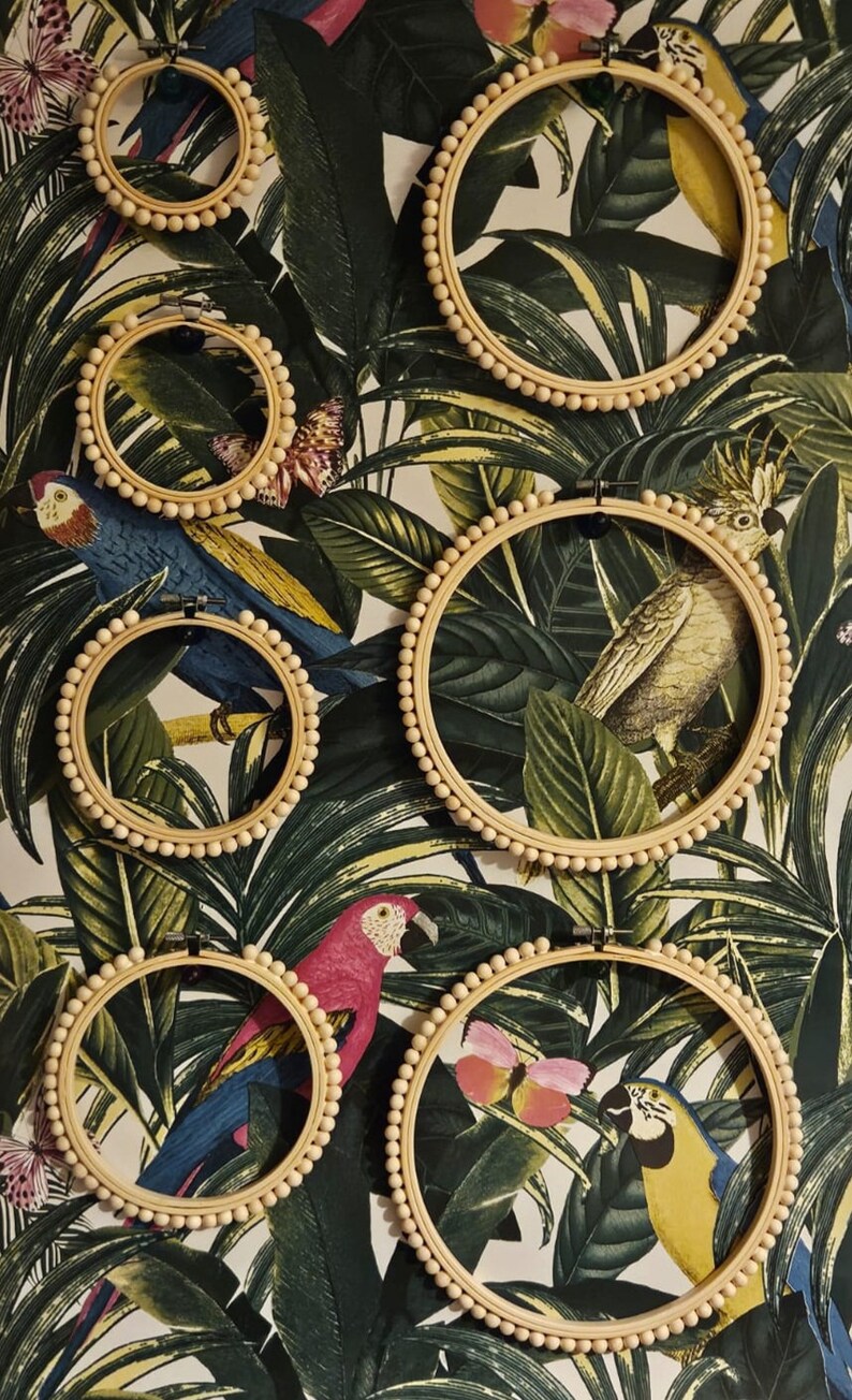 Puede incluir: Una colecci&oacute;n de aros de bordado de madera, cada uno adornado con peque&ntilde;as cuentas de color claro, se exhibe contra un vibrante papel pintado tropical. El papel pintado presenta exuberantes hojas verdes, loros coloridos y mariposas, creando una exhibici&oacute;n decorativa y art&iacute;stica.