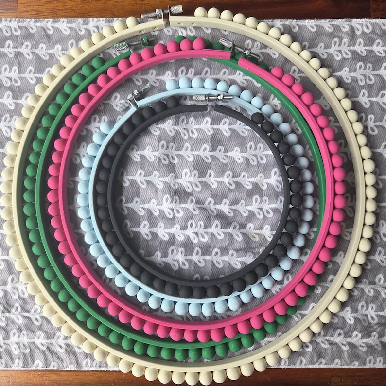 Puede incluir: Un conjunto de bastidores de bordado coloridos dispuestos en c&iacute;rculo. Los colores incluyen rosa, verde, azul y crema. Cada bastidor est&aacute; adornado con peque&ntilde;as bolas decorativas en el borde exterior. Se observan diferentes tama&ntilde;os.