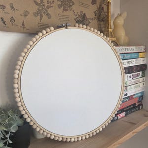Op de afbeelding: Een ronde borduurring met een wit canvas en een houten kralenrand. De ring wordt tentoongesteld op een houten plank, met boeken en decoratieve items. De tekst "fuschia" is zichtbaar.