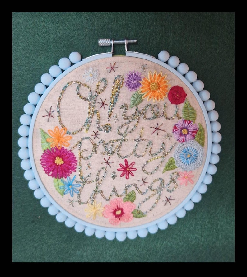 Puede incluir: Arte de aro bordado con la frase "Oh! You Pretty Things" en escritura cursiva, rodeada de coloridas flores bordadas y acentos de estrellas. El aro es azul claro con un ribete de pomp&oacute;n, sobre un fondo verde.