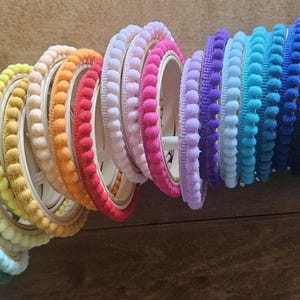 Pode incluir: Uma coleção de carretéis de guarnição de pompom colorido, dispostos em um padrão de arco-íris. A guarnição é feita de pompons macios e fofinhos e é perfeita para adicionar um toque de fantasia a qualquer projeto.