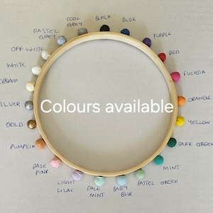 Puede incluir: Un bastidor de bordado de madera con pompones de colores colocados alrededor del borde. Los pompones son de varios colores, incluyendo azul, morado, rojo, naranja y verde. El texto central dice "Colores disponibles."
