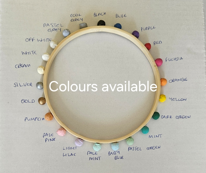 Puede incluir: Un bastidor de bordado de madera con pompones de colores colocados alrededor del borde. Los pompones son de varios colores, incluyendo azul, morado, rojo, naranja y verde. El texto central dice "Colores disponibles."