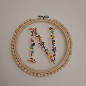 Può includere: Un telaio da ricamo in legno con bordo di perline contiene un tessuto bianco trasparente. La lettera "N" è formata con fiori secchi colorati. Il telaio è montato su una parete bianca, creando un monogramma floreale decorativo.