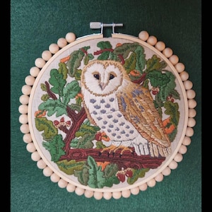 Puede incluir: Arte de aro de b&uacute;ho bordado. La obra de arte presenta una representaci&oacute;n detallada de un b&uacute;ho com&uacute;n posado en una rama, rodeado de follaje verde y bellotas. El b&uacute;ho tiene plumas blancas, marrones y grises. El bordado est&aacute; enmarcado en un aro de madera con cuentas de madera.