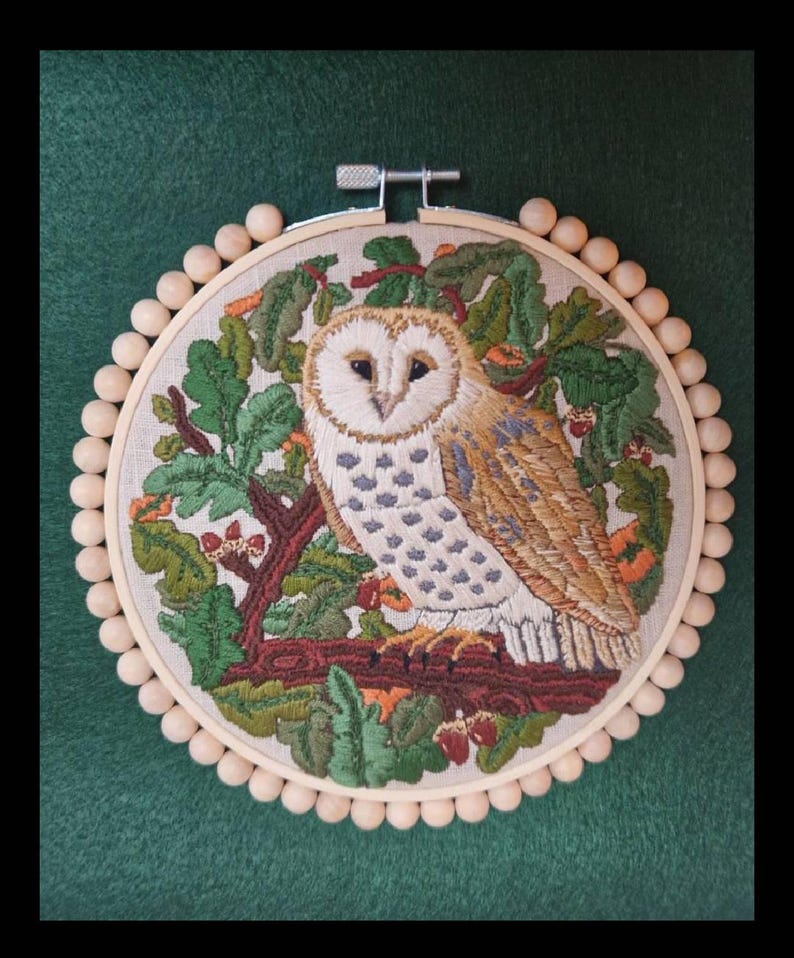 Puede incluir: Arte de aro de b&uacute;ho bordado. La obra de arte presenta una representaci&oacute;n detallada de un b&uacute;ho com&uacute;n posado en una rama, rodeado de follaje verde y bellotas. El b&uacute;ho tiene plumas blancas, marrones y grises. El bordado est&aacute; enmarcado en un aro de madera con cuentas de madera.
