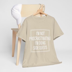 May include: A beige t-shirt with a white box and the text "I'm not procrastinating I'm doing side quests".