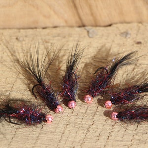 May include: Six handmade fishing flies. Each fly features a black body with red and iridescent accents, a pink bead head, and a curved hook. The flies are arranged on a wooden surface.