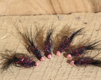 May include: Six fishing flies, handmade with black bodies, red and iridescent detailing, pink bead heads, and curved hooks. They are arranged on a wooden surface.