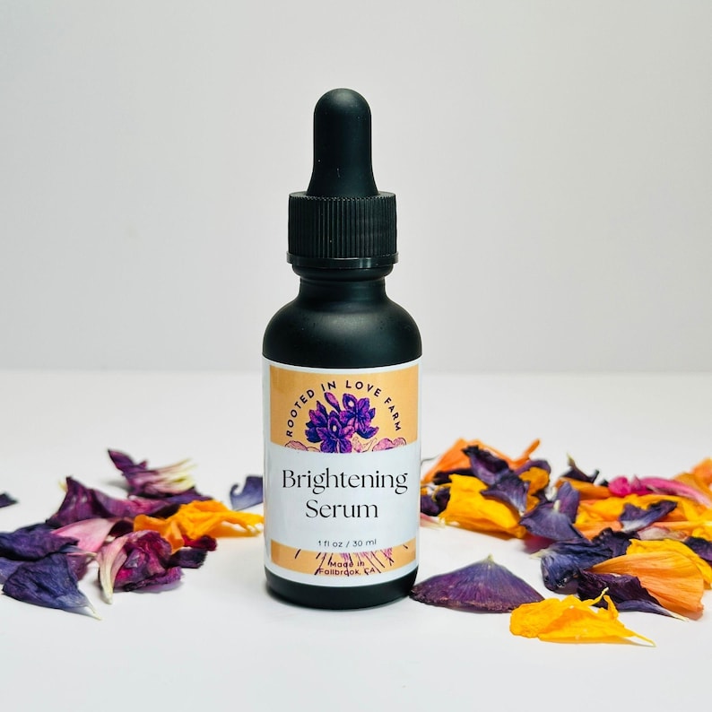 May include: A black glass bottle of Brightening Serum with a dropper, 1 fl oz / 30 ml. The label features text and a purple floral design. The bottle is surrounded by colorful flower petals. The product is from Rooted in Love Farm.