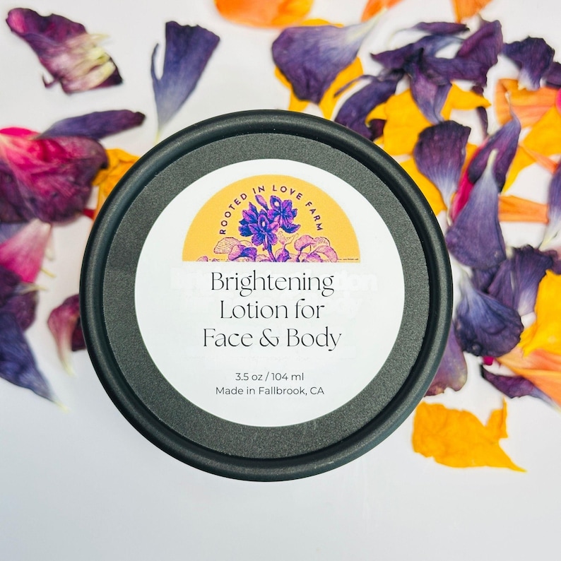 May include: A black round container of Brightening Lotion for Face & Body, 3.5 oz. The label features a floral design and the words "Rooted in Love Farm." The container is surrounded by colorful flower petals.