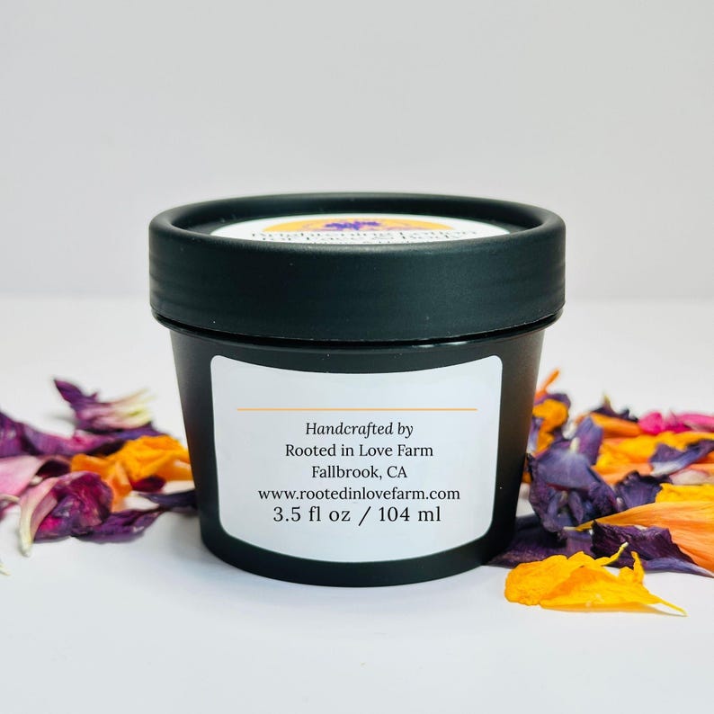 May include: A black container with a white label displaying text. The label reads "Handcrafted by Rooted in Love Farm Fallbrook, CA www.rootedinlovefarm.com 3.5 fl oz / 104 ml." The container is surrounded by colorful flower petals.