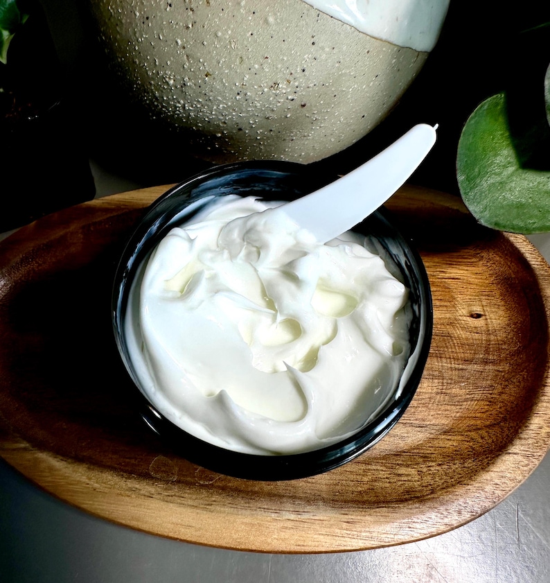 May include: A black glass bowl filled with white cream, with a white spatula resting inside. The bowl sits on a round, wooden tray. A ceramic pot and green plant are in the background.